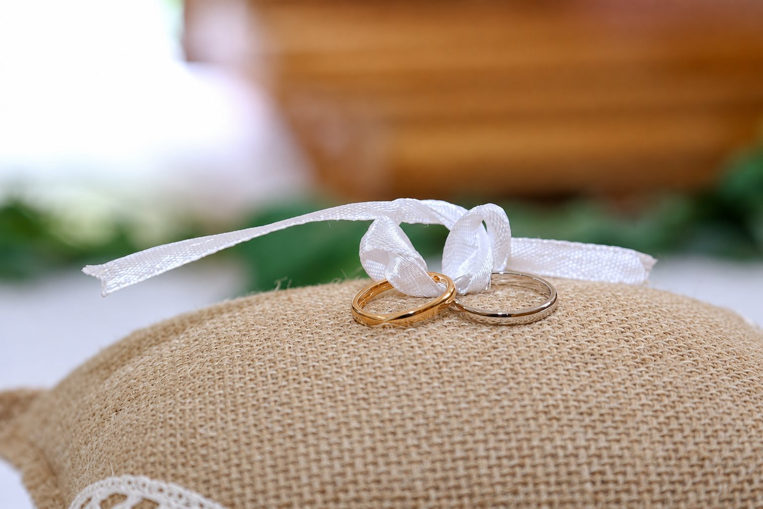 Deux alliances, l'une en or rose et l'autre en argent, sont attachées par un ruban de satin blanc sur un coussin en toile de jute.