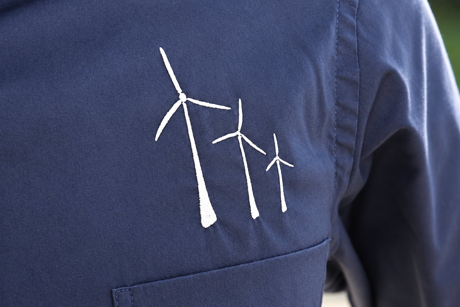 Gros plan sur une chemise bleu marine ornée d'une broderie blanche représentant trois éoliennes de tailles décroissantes.