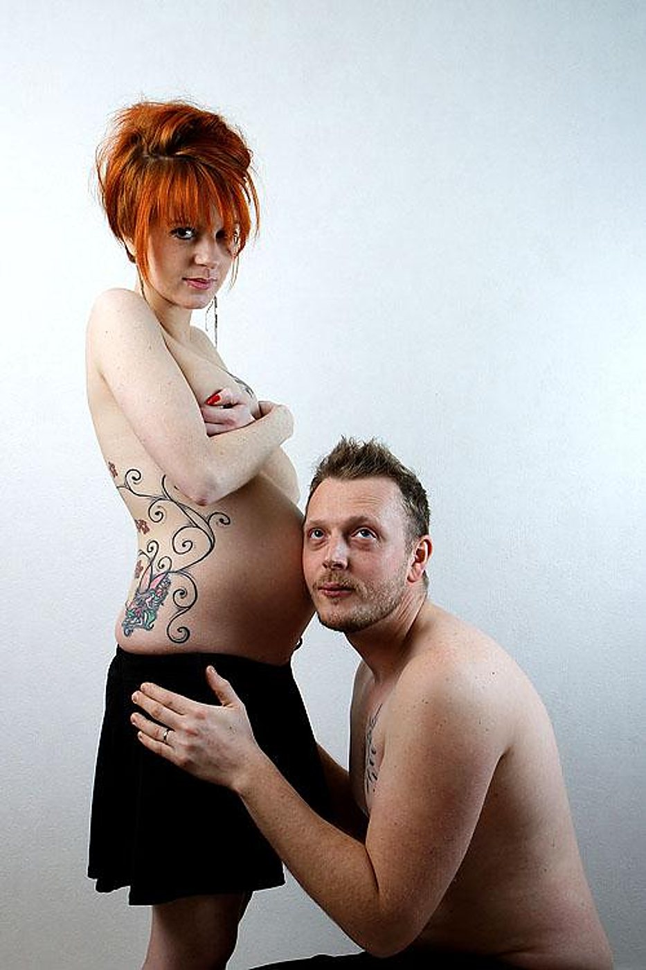 Une femme enceinte aux cheveux roux et tatouages se tient debout tandis qu'un homme agenouillé pose l'oreille contre son ventre, sur fond clair uni.