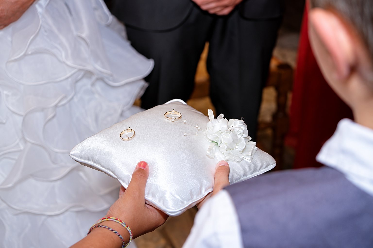 Un enfant tient un coussin de satin blanc orné de fleurs sur lequel reposent deux alliances, devant la robe de la mariée.