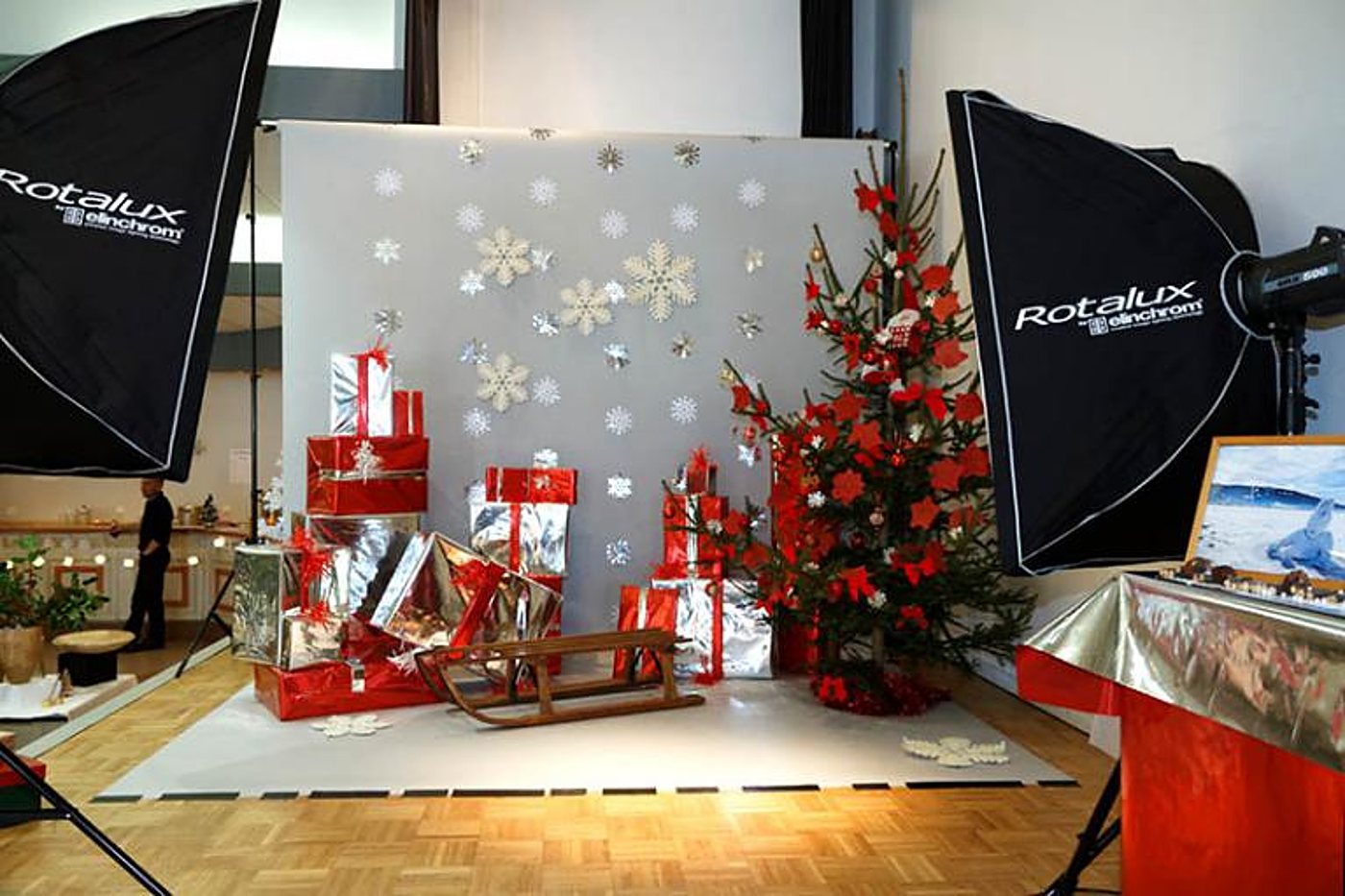 Un décor de Noël avec sapin rouge, cadeaux argentés et rouges, luge en bois et flocons sur fond gris, entouré de deux softboxes Rotalux.