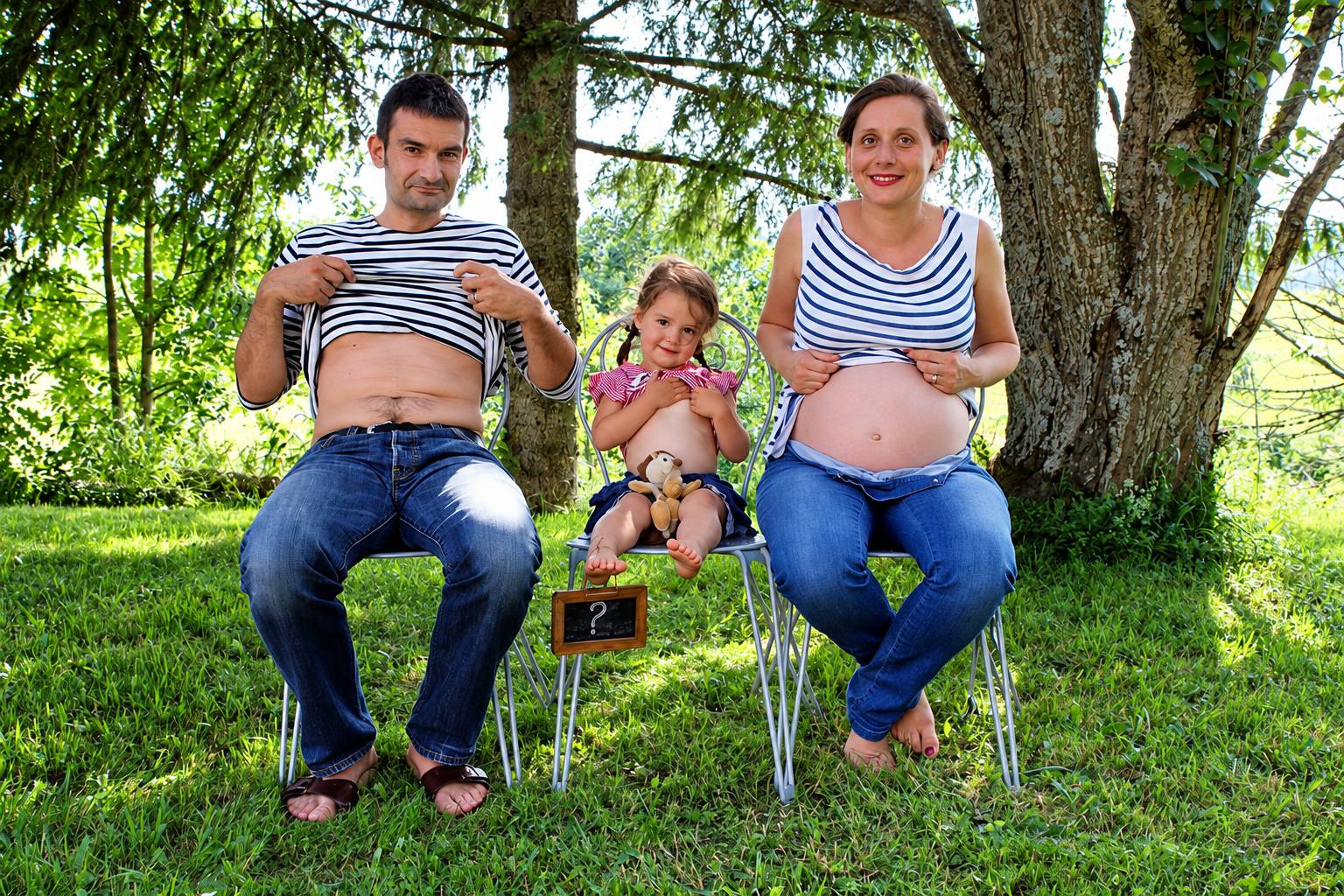 Un homme, une femme enceinte et une petite fille sont assis sur des chaises métalliques dans l'herbe sous les arbres, tous montrant leur ventre, avec une petite ardoise portant un point d'interrogation.