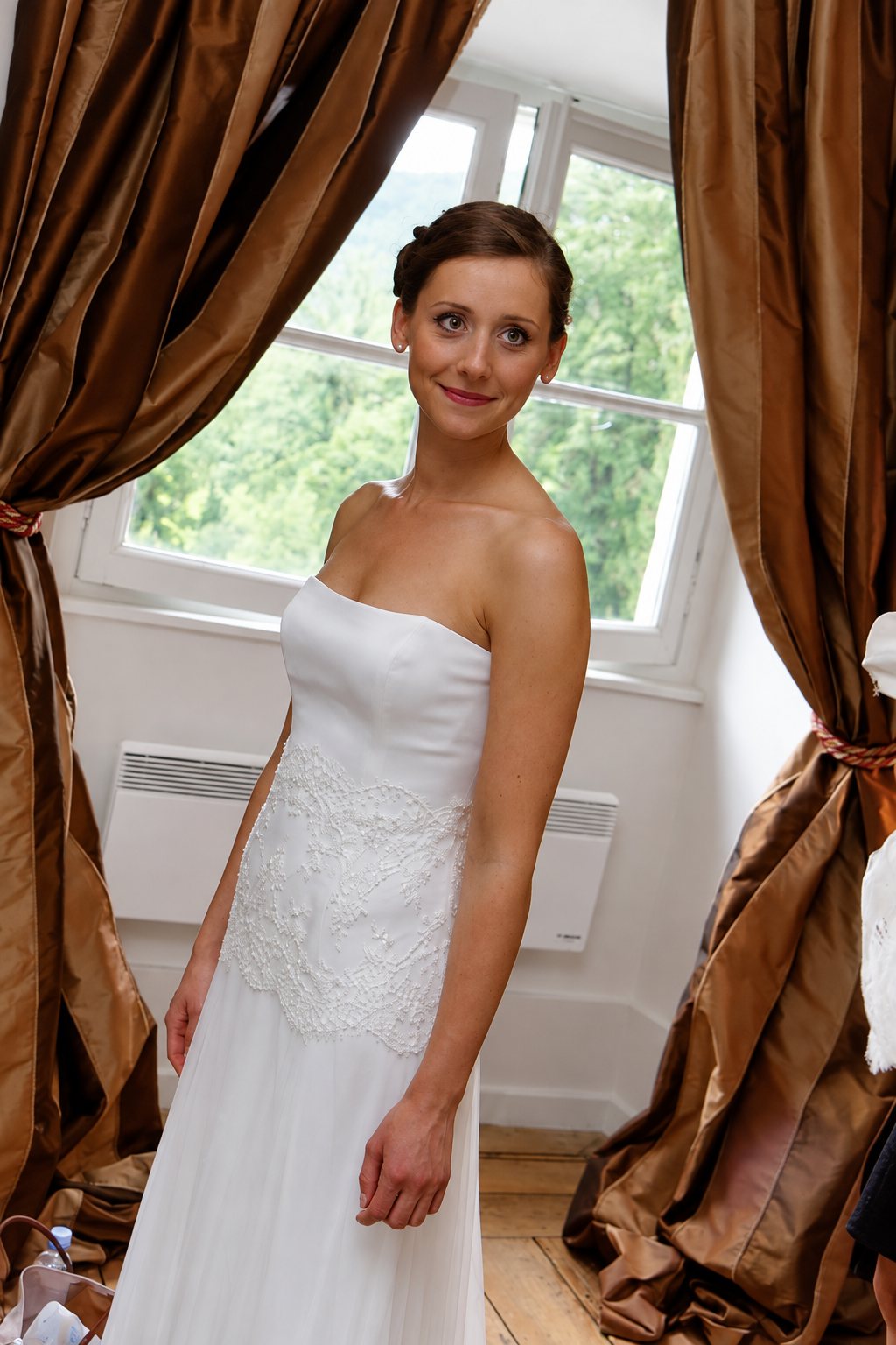 Une jeune femme souriante pose dans une robe de mariée bustier ornée de dentelle perlée, entre deux rideaux de soie cuivrés devant une fenêtre.