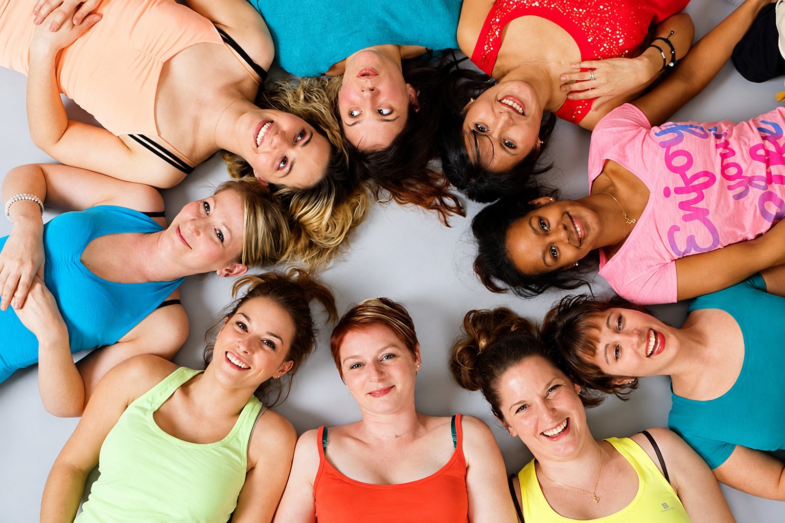 Huit femmes en tenues de sport colorées sont allongées sur le sol, têtes vers le centre, et sourient à l'objectif vu du dessus.