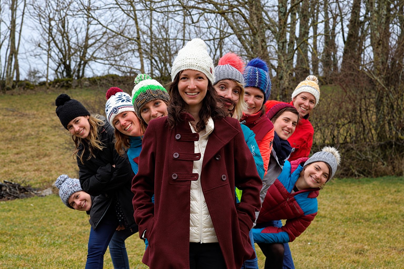 Un groupe de femmes souriantes en bonnets d'hiver colorés pose en file penchée derrière une femme en manteau bordeaux, sur une pelouse devant des arbres sans feuilles.
