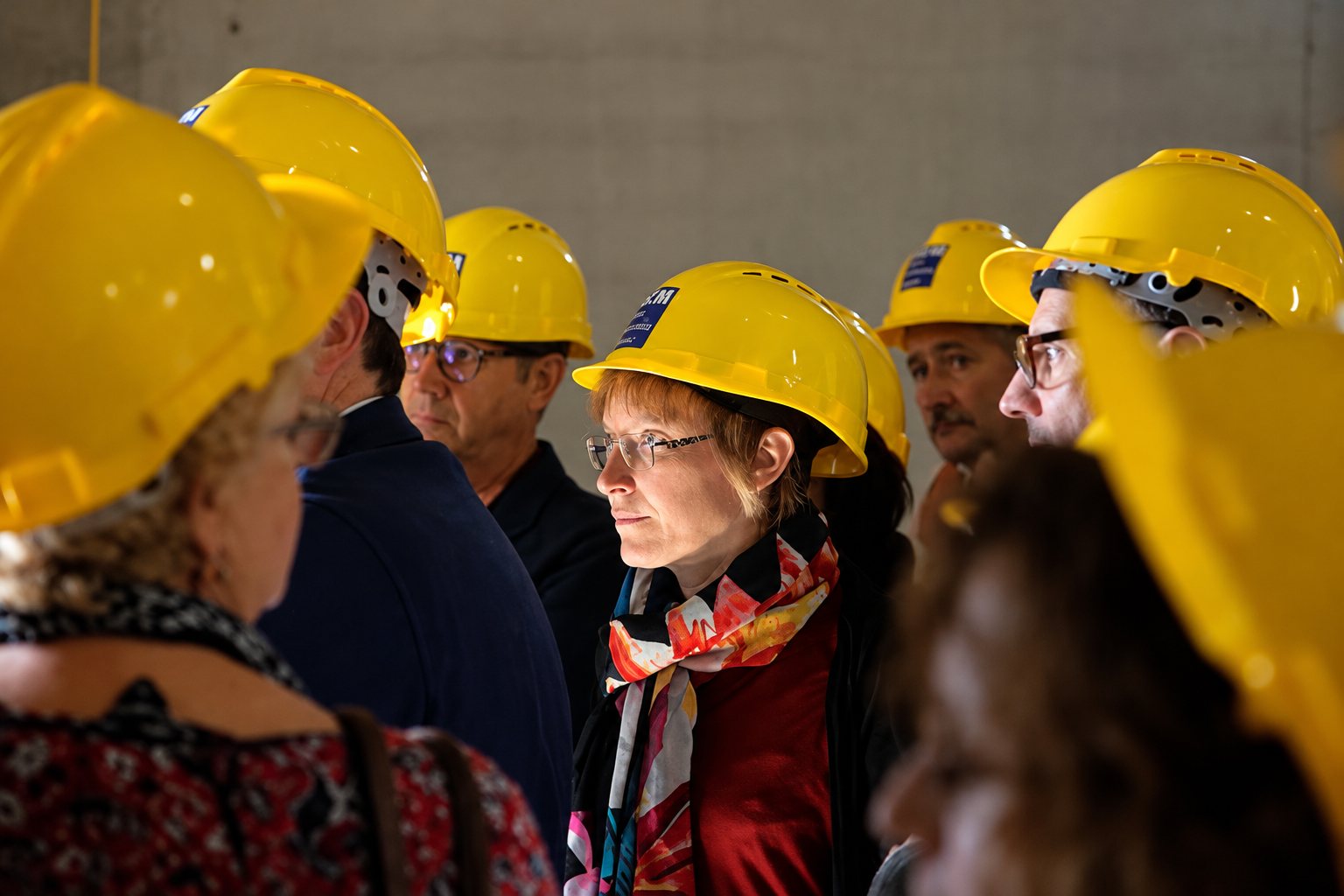 Un groupe de personnes portant des casques de chantier jaunes écoute attentivement une présentation à l'intérieur d'un bâtiment industriel sombre.
