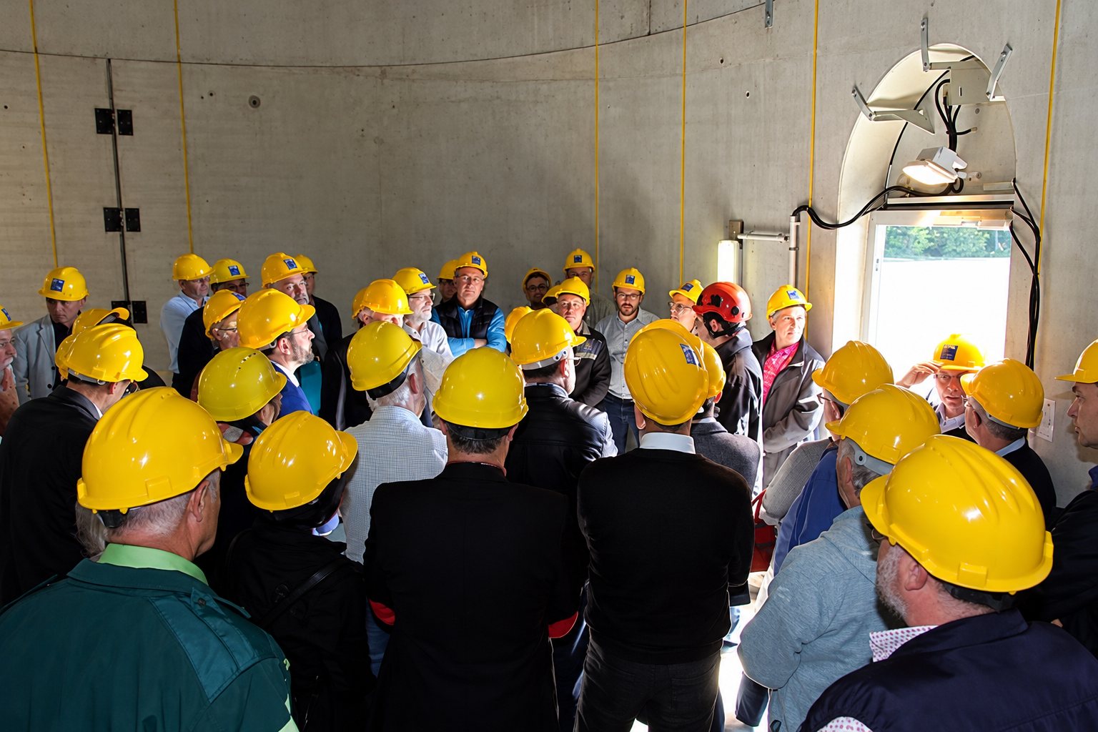 Un groupe de personnes portant des casques de chantier jaunes se rassemble dans une grande salle en béton, près d'une porte ouverte donnant sur l'extérieur.