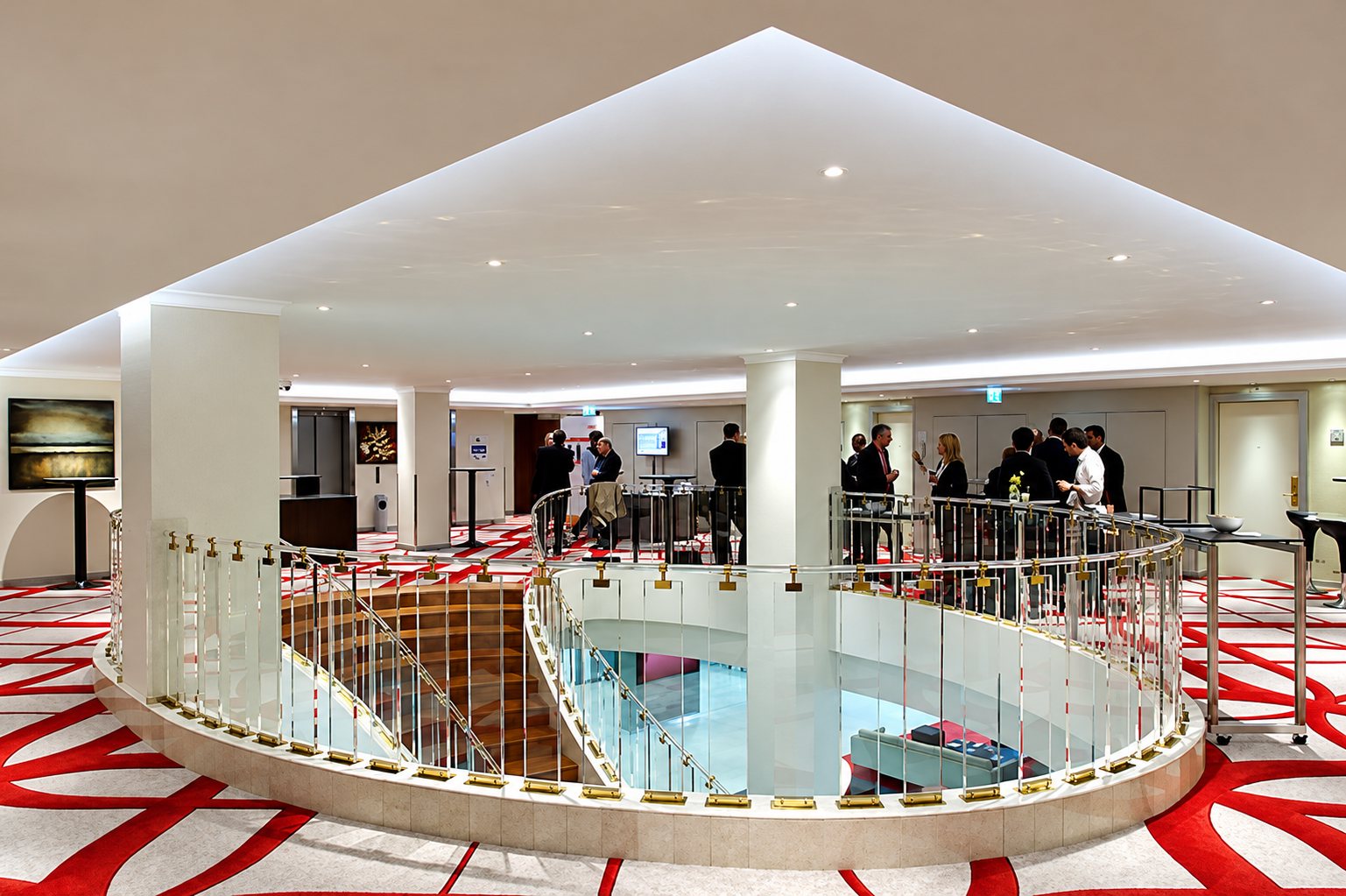 Un vaste hall d'hôtel au tapis rouge et blanc, avec une balustrade de verre autour d'une mezzanine et des personnes qui discutent au fond.
