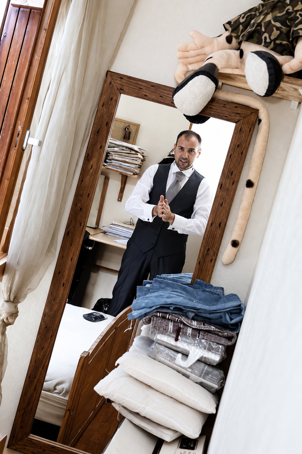 Photo en noir et blanc d'un homme en chemise, gilet et cravate qui se prépare, vu dans un miroir à cadre en bois rustique, avec des oreillers et du linge empilés au premier plan.