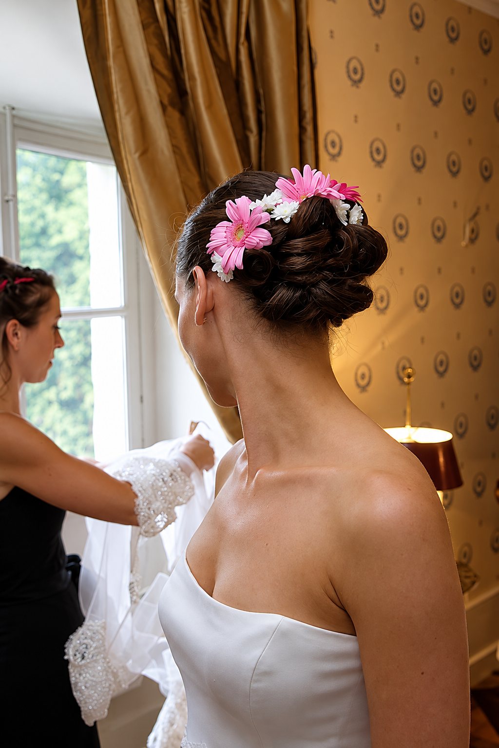 Une mariée en robe bustier blanc, vue de dos, porte un chignon décoré de gerberas roses et de petites fleurs blanches, tandis qu'une femme tient le voile près d'une fenêtre.
