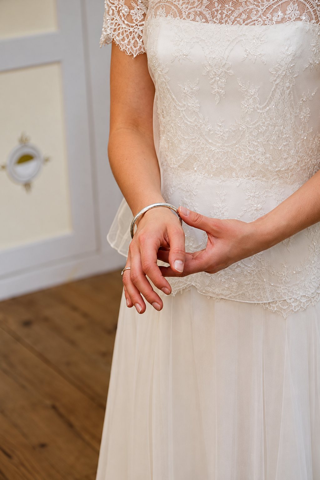 Gros plan sur les mains d'une mariée vêtue d'une robe en dentelle ivoire à manches courtes, un bracelet jonc argenté au poignet.