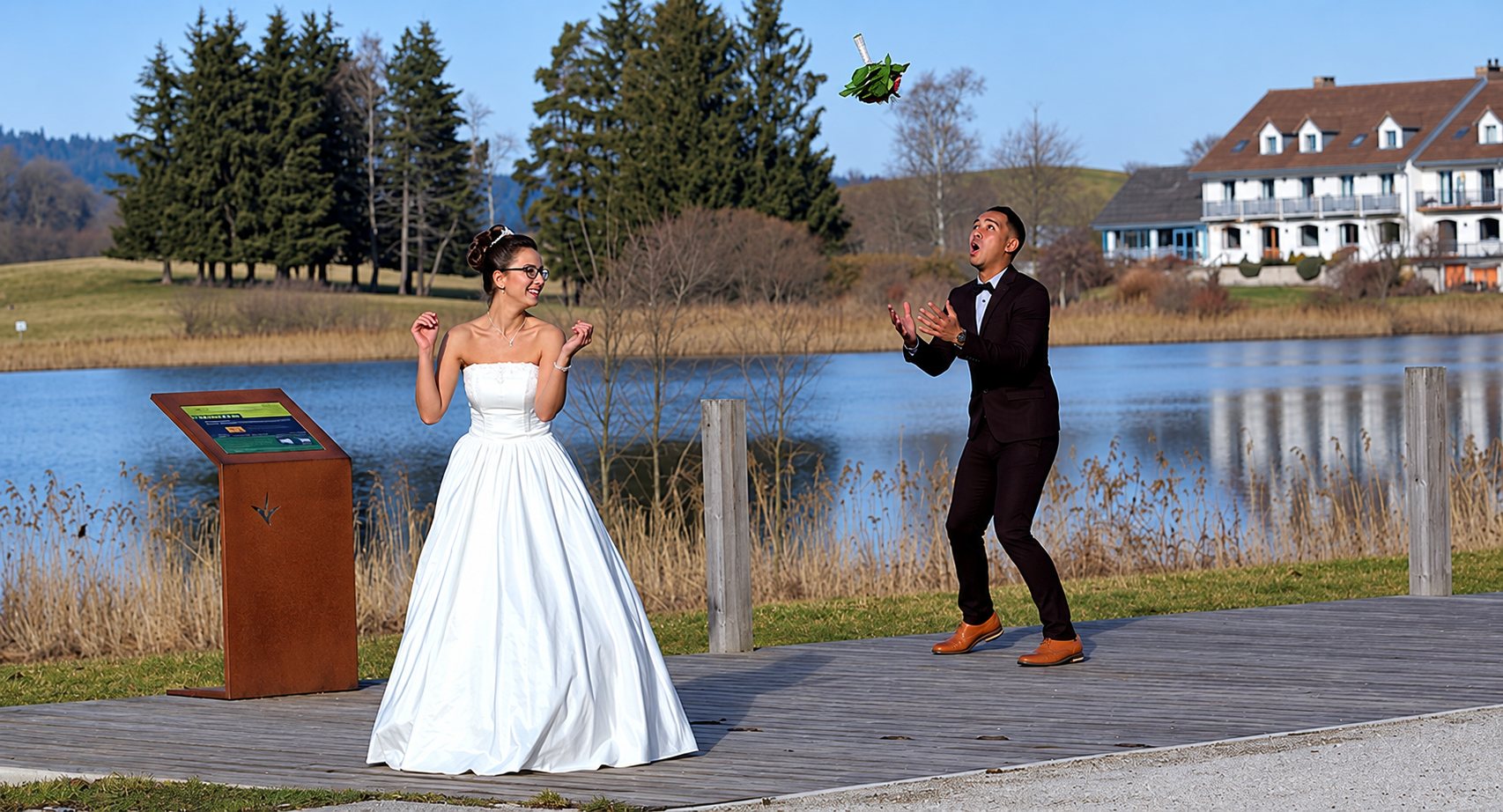 Une mariée en robe blanche regarde son marié rattraper un bouquet lancé en l'air, sur un ponton en bois au bord d'un lac, avec un grand bâtiment en arrière-plan.