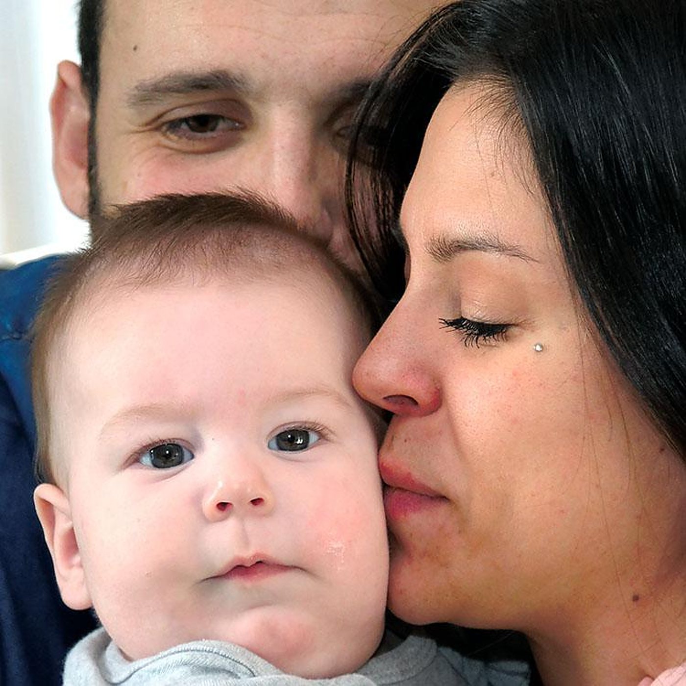 Gros plan d'un bébé aux yeux sombres, embrassé sur la joue par sa mère, avec le père visible derrière eux.