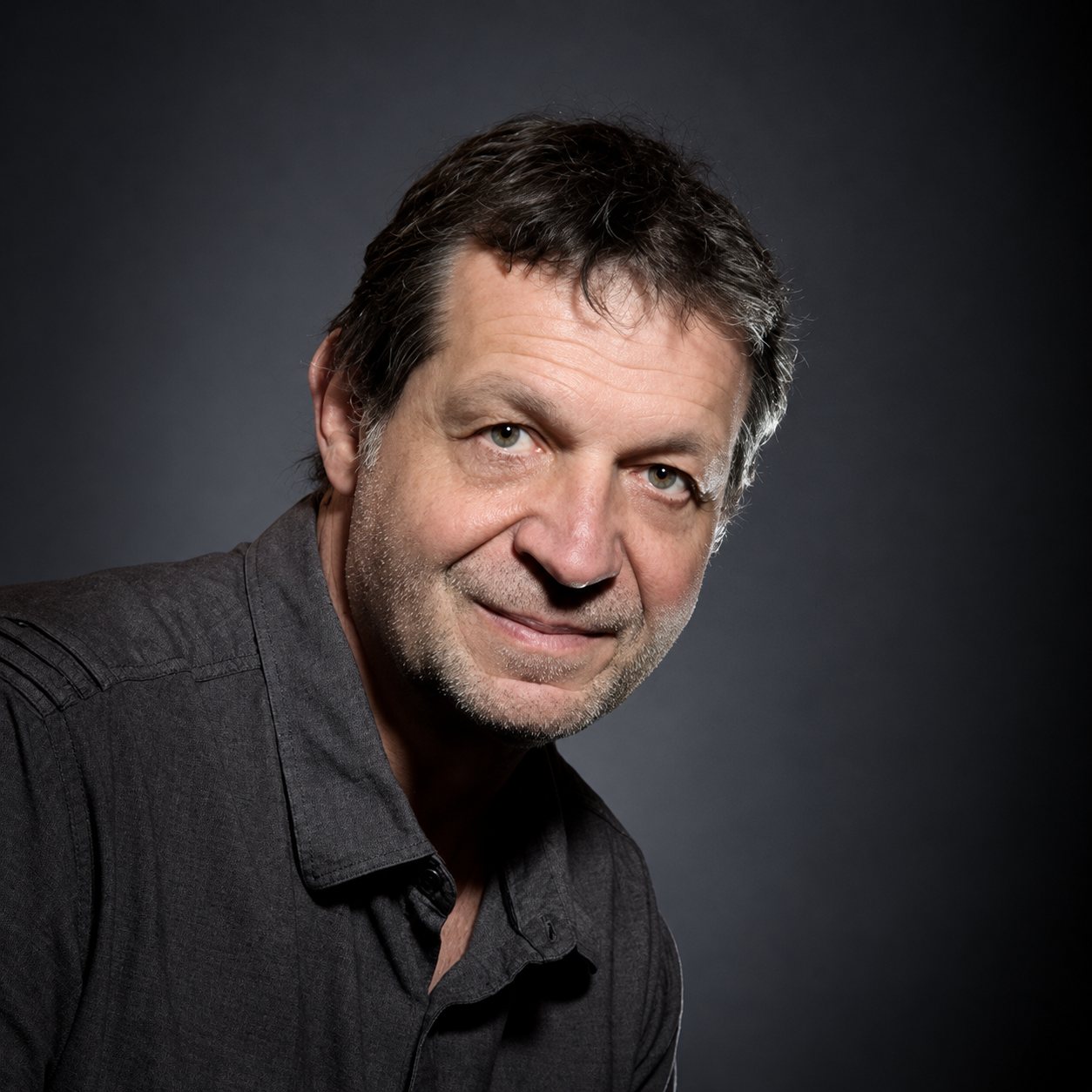 Portrait en studio d’un homme souriant aux cheveux bruns, sur fond sombre.