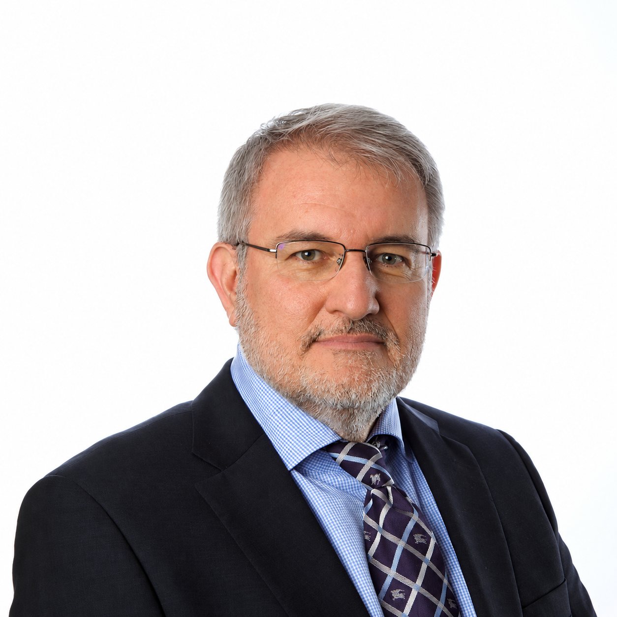 Portrait d'un homme aux cheveux gris, barbe courte et lunettes, vêtu d'un costume sombre, d'une chemise bleue à carreaux et d'une cravate violette, devant un fond blanc.
