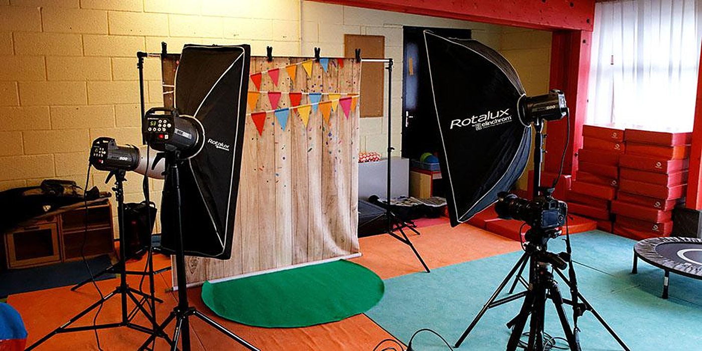 Un studio photo installé dans une salle colorée, avec un fond en bois décoré de fanions, deux softboxes Rotalux, des flashs sur pieds et un appareil photo sur trépied.