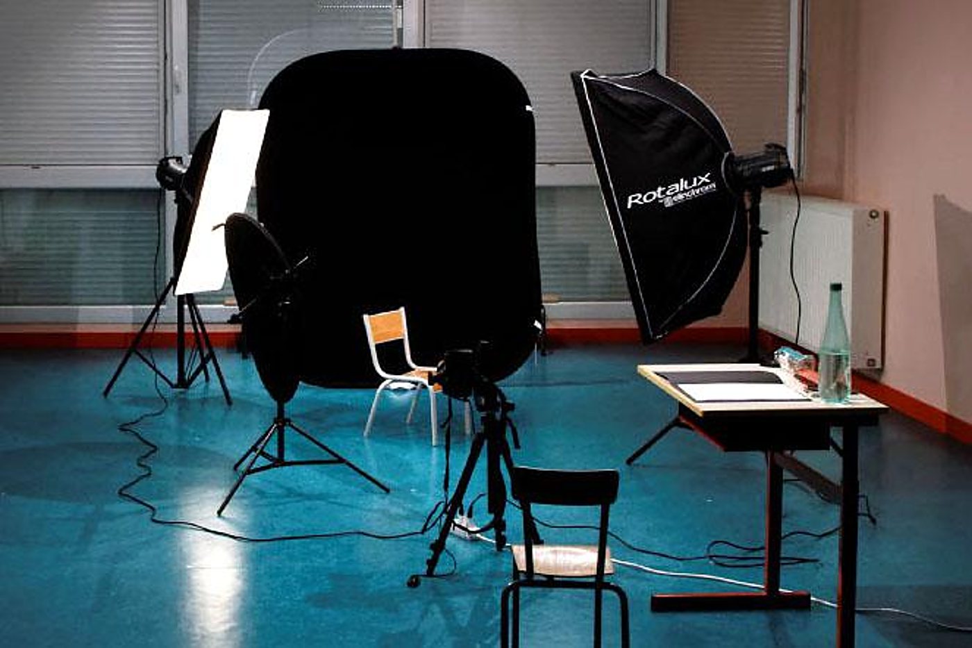 Matériel de studio photo installé dans une salle au sol bleu, avec softbox Rotalux, réflecteur noir, trépieds, chaises et table d'écolier.