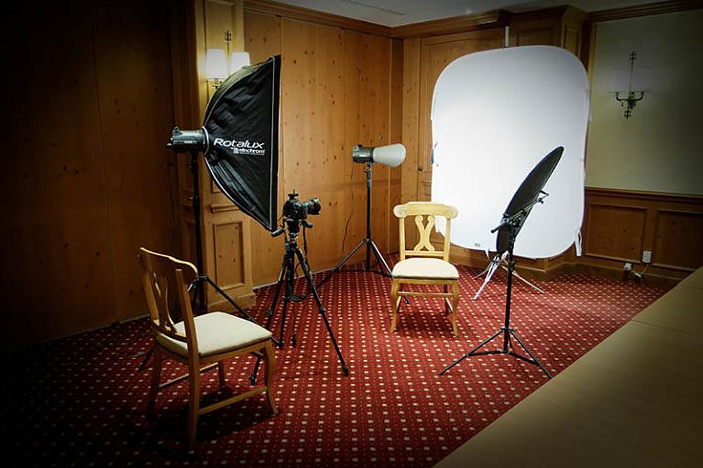 Un studio photo installé dans une pièce lambrissée de bois, avec softbox Rotalux, flashs sur pieds, appareil photo sur trépied, fond blanc et deux chaises sur un tapis rouge.