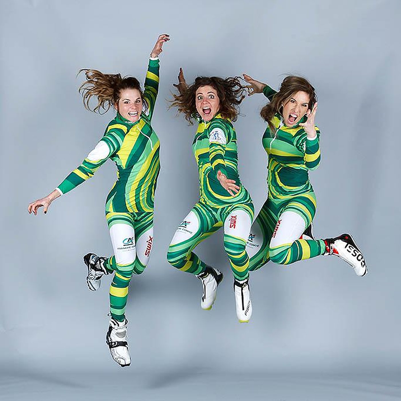 Trois femmes en combinaison de ski de fond verte, jaune et blanche sautent joyeusement devant un fond gris uni.